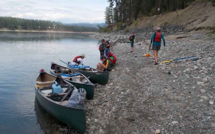three canoes are beached on a shore of a calm lake. a few students carry gear away from the canoes. 